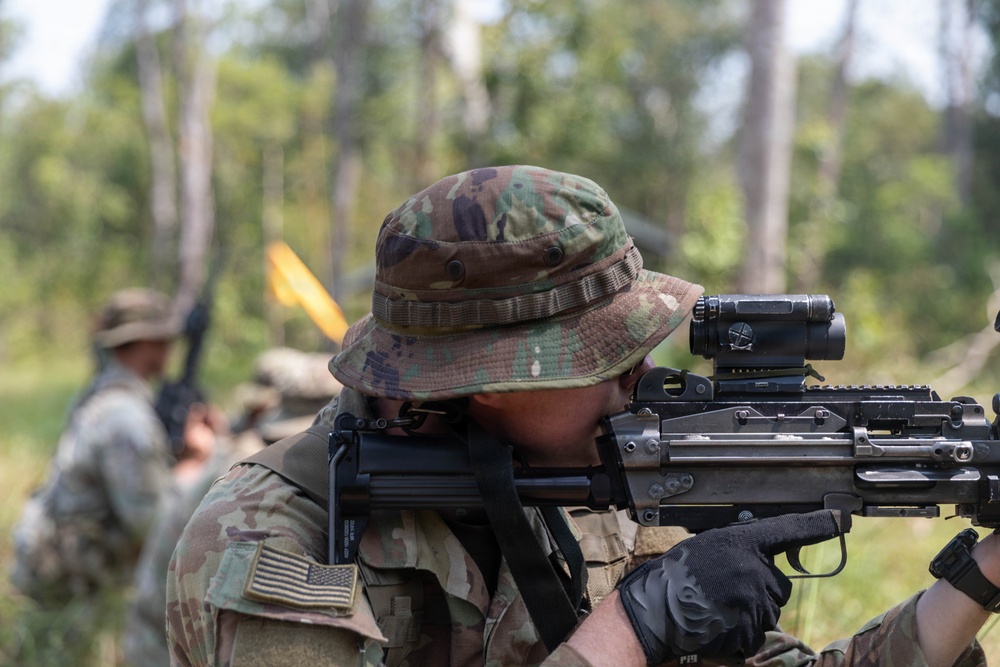 A U.S. Army soldier in MultiCam/OCP camouflage uniform is aiming a rifle equipped with an optic sight during what appears to be a field training exercise. The soldier is wearing a boonie hat and tactical gloves, with a U.S. flag patch visible on the left shoulder. Additional soldiers in the background suggest a group training scenario in a wooded outdoor environment.
