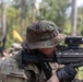 A U.S. Army soldier in MultiCam/OCP camouflage uniform is aiming a rifle equipped with an optic sight during what appears to be a field training exercise. The soldier is wearing a boonie hat and tactical gloves, with a U.S. flag patch visible on the left shoulder. Additional soldiers in the background suggest a group training scenario in a wooded outdoor environment.