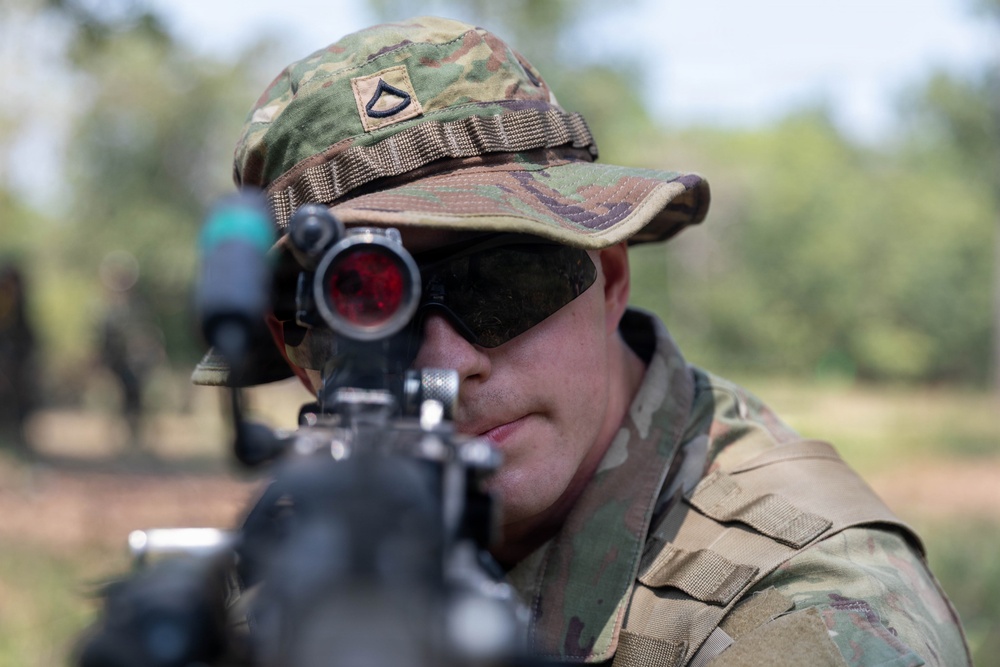 A U.S. Army soldier in Operational Camouflage Pattern (OCP) uniform is aiming a weapon directly toward the camera. The soldier wears a rank insignia patch on their patrol cap consistent with Private First Class (PFC). The image is a close-up shot taken outdoors in a wooded or field training environment.