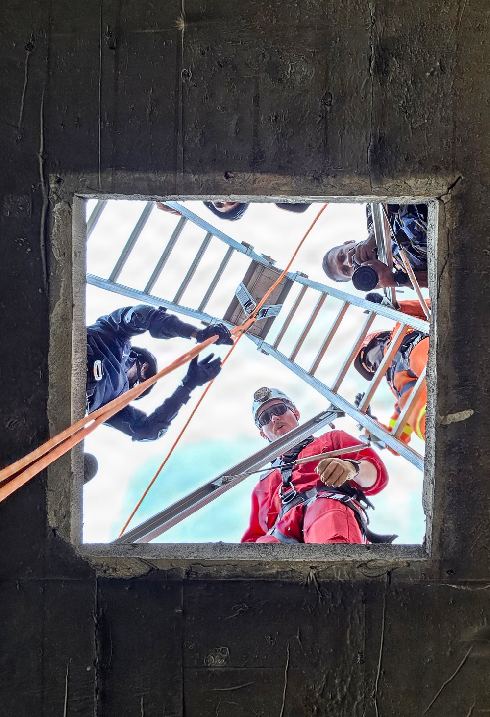 The image is taken from below looking up through a square concrete opening or hatch, showing multiple workers gathered around the opening from above. The workers are equipped with safety gear including harnesses, helmets, and gloves, and are managing orange ropes/lines along with an aluminum ladder positioned over the opening. This appears to be a confined space rescue or rope access operation being conducted by a trained team.