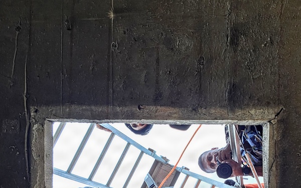 The image is taken from below looking up through a square concrete opening or hatch, showing multiple workers gathered around the opening from above. The workers are equipped with safety gear including harnesses, helmets, and gloves, and are managing orange ropes/lines along with an aluminum ladder positioned over the opening. This appears to be a confined space rescue or rope access operation being conducted by a trained team.