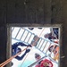 The image is taken from below looking up through a square concrete opening or hatch, showing multiple workers gathered around the opening from above. The workers are equipped with safety gear including harnesses, helmets, and gloves, and are managing orange ropes/lines along with an aluminum ladder positioned over the opening. This appears to be a confined space rescue or rope access operation being conducted by a trained team.