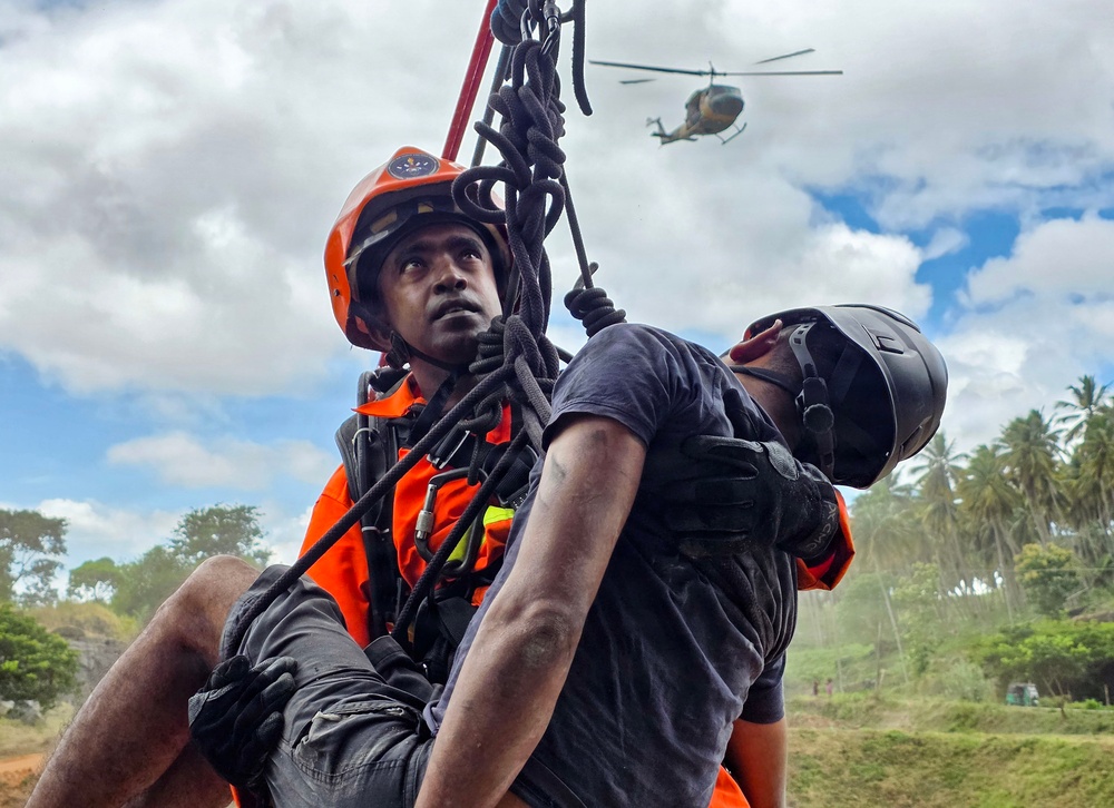 A search and rescue operation is underway, showing a rescuer in orange safety gear and helmet hoisting or supporting a casualty/victim using ropes and rigging equipment. A helicopter is visible in the background sky, suggesting an aerial component to the rescue operation. The scene appears to take place in a tropical environment based on the palm trees and lush vegetation visible in the background.