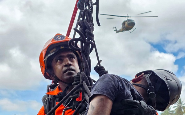 A search and rescue operation is underway, showing a rescuer in orange safety gear and helmet hoisting or supporting a casualty/victim using ropes and rigging equipment. A helicopter is visible in the background sky, suggesting an aerial component to the rescue operation. The scene appears to take place in a tropical environment based on the palm trees and lush vegetation visible in the background.