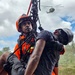 A search and rescue operation is underway, showing a rescuer in orange safety gear and helmet hoisting or supporting a casualty/victim using ropes and rigging equipment. A helicopter is visible in the background sky, suggesting an aerial component to the rescue operation. The scene appears to take place in a tropical environment based on the palm trees and lush vegetation visible in the background.