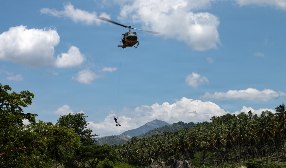 A military or utility helicopter, consistent with a Bell UH-1 'Huey' design, hovers over a tropical jungle landscape while a person dangles from a rope or hoist line beneath it. The scene appears to depict a hoist rescue, rappelling, or extraction operation. The lush tropical vegetation, palm trees, and mountainous background suggest a Southeast Asian or Caribbean setting.