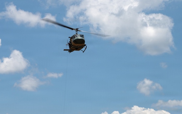 A military or utility helicopter, consistent with a Bell UH-1 'Huey' design, hovers over a tropical jungle landscape while a person dangles from a rope or hoist line beneath it. The scene appears to depict a hoist rescue, rappelling, or extraction operation. The lush tropical vegetation, palm trees, and mountainous background suggest a Southeast Asian or Caribbean setting.