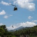 A military or utility helicopter, consistent with a Bell UH-1 'Huey' design, hovers over a tropical jungle landscape while a person dangles from a rope or hoist line beneath it. The scene appears to depict a hoist rescue, rappelling, or extraction operation. The lush tropical vegetation, palm trees, and mountainous background suggest a Southeast Asian or Caribbean setting.