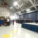 A formal military ceremony is taking place inside a large aircraft hangar. A senior officer in dress uniform is saluting on a raised stage, flanked by flags including the American flag and what appears to be an Air Force flag. A large audience of military personnel and civilians is assembled to the left observing the ceremony.