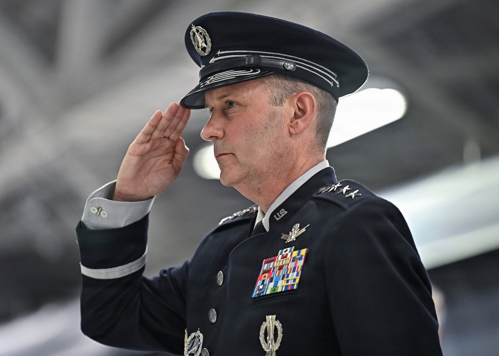 A senior U.S. military officer in dress uniform is rendering a formal salute. The individual wears a dark blue service dress uniform consistent with the U.S. Air Force or U.S. Space Force, adorned with multiple ribbons, badges, and what appear to be four-star rank insignia on the shoulder. The setting appears to be an aircraft hangar or large military facility.