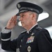 A senior U.S. military officer in dress uniform is rendering a formal salute. The individual wears a dark blue service dress uniform consistent with the U.S. Air Force or U.S. Space Force, adorned with multiple ribbons, badges, and what appear to be four-star rank insignia on the shoulder. The setting appears to be an aircraft hangar or large military facility.