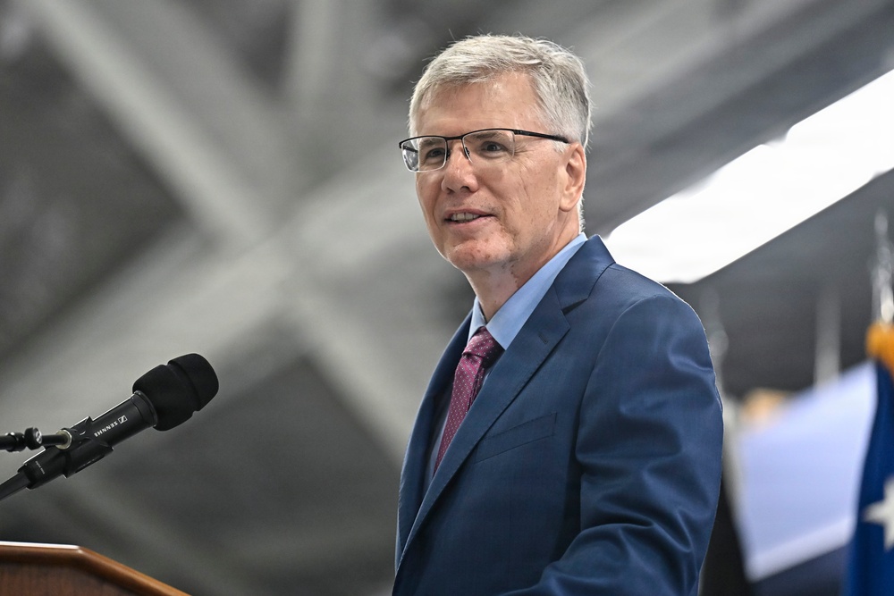 A middle-aged man with gray hair and rectangular glasses is speaking at a podium equipped with a Sennheiser microphone. He is dressed in a navy blue suit with a light blue dress shirt and a burgundy/maroon patterned tie. The setting appears to be an indoor venue, possibly a hangar or large facility, with a flag partially visible in the background.