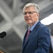 A middle-aged man with gray hair and rectangular glasses is speaking at a podium equipped with a Sennheiser microphone. He is dressed in a navy blue suit with a light blue dress shirt and a burgundy/maroon patterned tie. The setting appears to be an indoor venue, possibly a hangar or large facility, with a flag partially visible in the background.