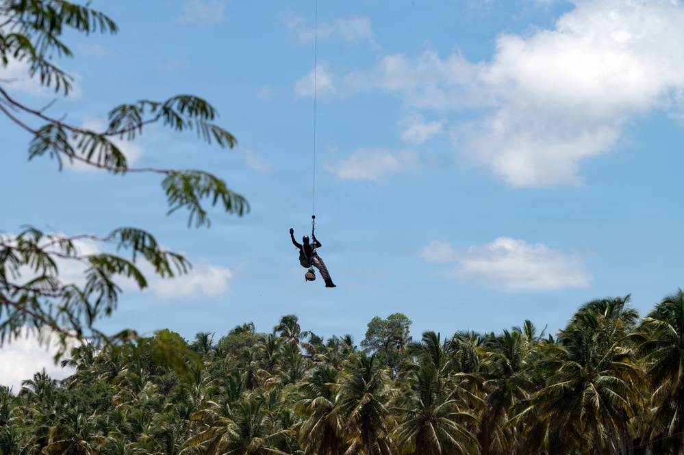 A person is suspended in mid-air from a single rope or cable against a partly cloudy blue sky. The silhouetted figure appears to be dangling or being lifted, with what looks like a bag or bundle attached below them. A dense canopy of tropical palm trees forms the background horizon.