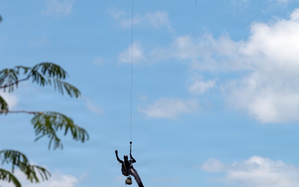 A person is suspended in mid-air from a single rope or cable against a partly cloudy blue sky. The silhouetted figure appears to be dangling or being lifted, with what looks like a bag or bundle attached below them. A dense canopy of tropical palm trees forms the background horizon.