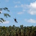 A person is suspended in mid-air from a single rope or cable against a partly cloudy blue sky. The silhouetted figure appears to be dangling or being lifted, with what looks like a bag or bundle attached below them. A dense canopy of tropical palm trees forms the background horizon.