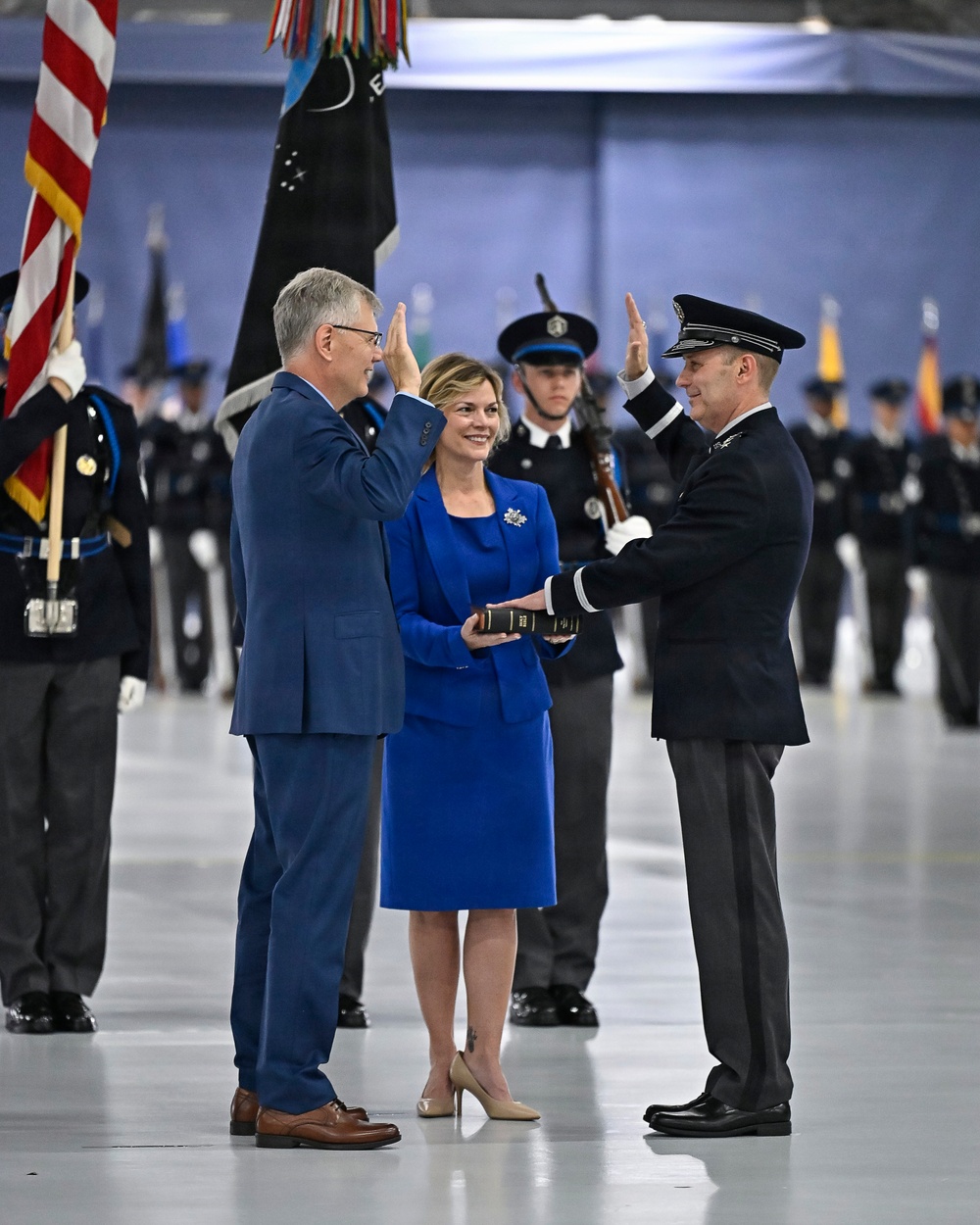 A formal military or government swearing-in ceremony taking place inside a large hangar or indoor venue. A man in a blue civilian suit is administering an oath to a uniformed officer in dark dress uniform, while a woman in a royal blue dress holds a book (likely a Bible) between them. Honor guard members and color bearers with flags, including the U.S. flag and what appears to be a Space Force flag, stand in formation in the background.