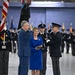 A formal military or government swearing-in ceremony taking place inside a large hangar or indoor venue. A man in a blue civilian suit is administering an oath to a uniformed officer in dark dress uniform, while a woman in a royal blue dress holds a book (likely a Bible) between them. Honor guard members and color bearers with flags, including the U.S. flag and what appears to be a Space Force flag, stand in formation in the background.