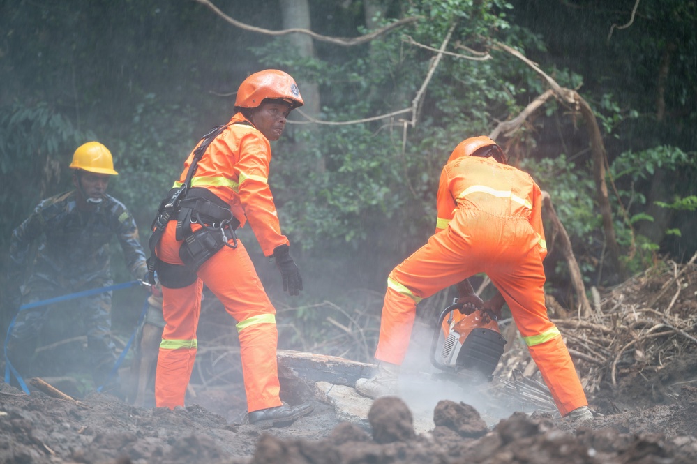 Emergency rescue workers in orange high-visibility uniforms and helmets are conducting operations at what appears to be a landslide or disaster site. Two workers in orange suits are prominently featured, with one operating a chainsaw to cut through debris while the other observes. A third person in blue military or utility clothing with a yellow hard hat is visible in the background.
