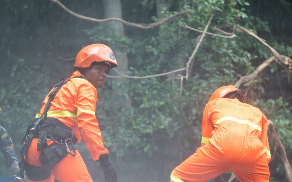 Emergency rescue workers in orange high-visibility uniforms and helmets are conducting operations at what appears to be a landslide or disaster site. Two workers in orange suits are prominently featured, with one operating a chainsaw to cut through debris while the other observes. A third person in blue military or utility clothing with a yellow hard hat is visible in the background.