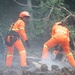 Emergency rescue workers in orange high-visibility uniforms and helmets are conducting operations at what appears to be a landslide or disaster site. Two workers in orange suits are prominently featured, with one operating a chainsaw to cut through debris while the other observes. A third person in blue military or utility clothing with a yellow hard hat is visible in the background.