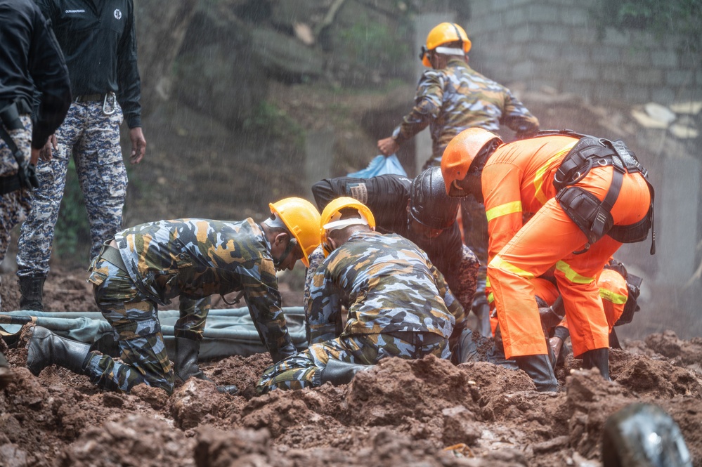 Rescue workers in camouflage uniforms and orange high-visibility suits are conducting search and rescue operations in muddy terrain, likely following a landslide. Multiple personnel wearing yellow hard hats are working together, some kneeling in the mud while handling what appears to be a body bag or stretcher. The operation is taking place in wet, rainy conditions with significant mud displacement visible.