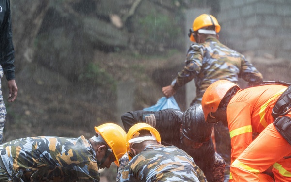 Rescue workers in camouflage uniforms and orange high-visibility suits are conducting search and rescue operations in muddy terrain, likely following a landslide. Multiple personnel wearing yellow hard hats are working together, some kneeling in the mud while handling what appears to be a body bag or stretcher. The operation is taking place in wet, rainy conditions with significant mud displacement visible.