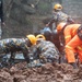 Rescue workers in camouflage uniforms and orange high-visibility suits are conducting search and rescue operations in muddy terrain, likely following a landslide. Multiple personnel wearing yellow hard hats are working together, some kneeling in the mud while handling what appears to be a body bag or stretcher. The operation is taking place in wet, rainy conditions with significant mud displacement visible.