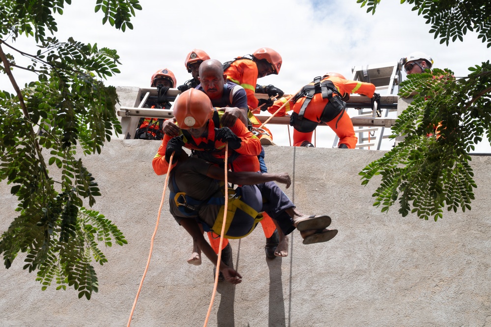 A rescue operation or training exercise is underway, with emergency responders in orange uniforms and helmets rappelling down a concrete wall while carrying a civilian. Multiple rescuers are visible at the top of the wall assisting with ropes and a ladder. The scene appears to be a vertical evacuation drill or live rescue scenario.