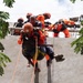 A rescue operation or training exercise is underway, with emergency responders in orange uniforms and helmets rappelling down a concrete wall while carrying a civilian. Multiple rescuers are visible at the top of the wall assisting with ropes and a ladder. The scene appears to be a vertical evacuation drill or live rescue scenario.