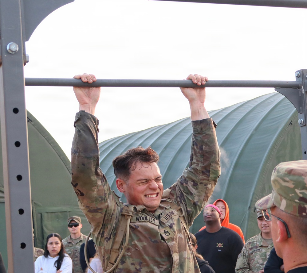 A U.S. Army soldier named Woolley, identifiable by the name tape on their uniform, is performing pull-ups on a metal bar while grimacing with effort. The soldier is wearing full Army Combat Uniform (ACU) in OCP (Operational Camouflage Pattern) and appears to be participating in a physical fitness test or competition. A crowd of military personnel and civilians observes in the background near green military tent structures.