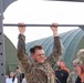A U.S. Army soldier named Woolley, identifiable by the name tape on their uniform, is performing pull-ups on a metal bar while grimacing with effort. The soldier is wearing full Army Combat Uniform (ACU) in OCP (Operational Camouflage Pattern) and appears to be participating in a physical fitness test or competition. A crowd of military personnel and civilians observes in the background near green military tent structures.