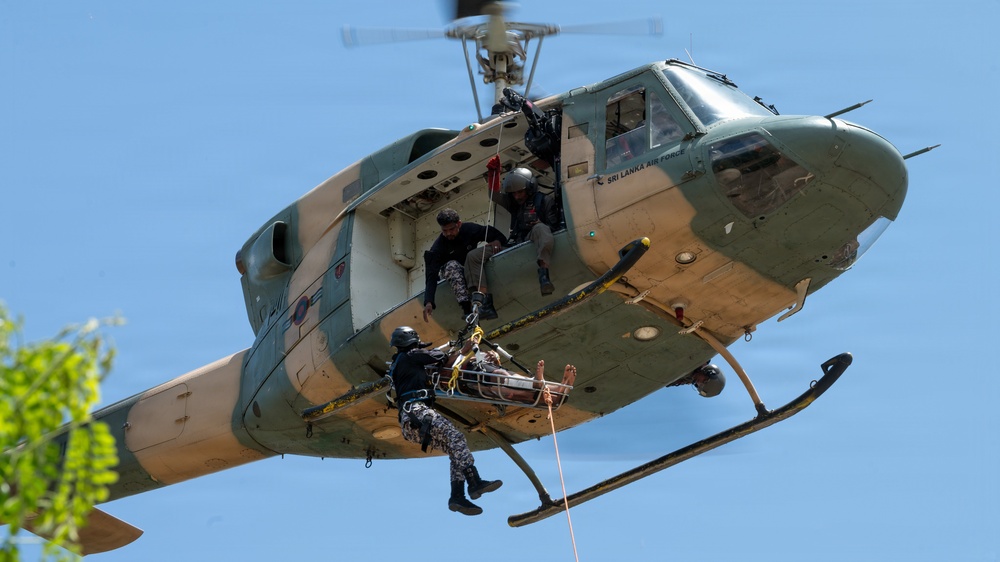 A Sri Lanka Air Force helicopter is conducting a medical evacuation (MEDEVAC) or search-and-rescue operation. Personnel are hoisting a casualty on a stretcher/litter via rope while a crew member leans out of the open door to assist. This appears to be either a live rescue operation or a training exercise demonstration.