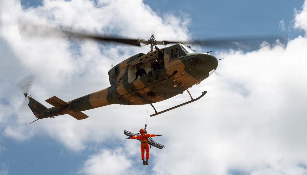 A military-style helicopter is performing a hoist rescue operation, with a person in an orange flight suit being lifted via a cable while holding a rescue stretcher/litter. The helicopter appears to be a Bell UH-1 'Huey' variant with an olive drab and tan camouflage paint scheme. Crew members are visible in the open side door of the helicopter overseeing the operation.