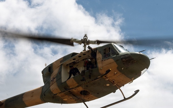 A military-style helicopter is performing a hoist rescue operation, with a person in an orange flight suit being lifted via a cable while holding a rescue stretcher/litter. The helicopter appears to be a Bell UH-1 'Huey' variant with an olive drab and tan camouflage paint scheme. Crew members are visible in the open side door of the helicopter overseeing the operation.