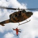 A military-style helicopter is performing a hoist rescue operation, with a person in an orange flight suit being lifted via a cable while holding a rescue stretcher/litter. The helicopter appears to be a Bell UH-1 'Huey' variant with an olive drab and tan camouflage paint scheme. Crew members are visible in the open side door of the helicopter overseeing the operation.