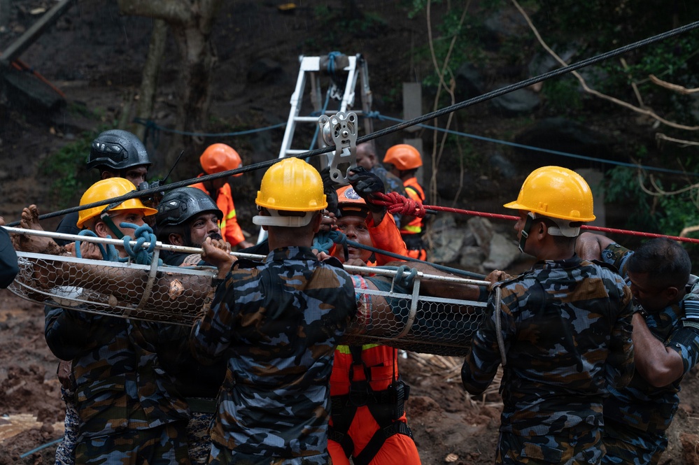 A search and rescue operation is underway at what appears to be a landslide or disaster site. Multiple rescue personnel in camouflage uniforms and hard hats are carrying a wire mesh stretcher (Stokes basket) with a victim, while a rope and pulley system is visible in the background. The muddy terrain and debris suggest a recent natural disaster such as a landslide or building collapse.