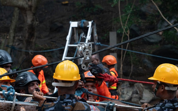 A search and rescue operation is underway at what appears to be a landslide or disaster site. Multiple rescue personnel in camouflage uniforms and hard hats are carrying a wire mesh stretcher (Stokes basket) with a victim, while a rope and pulley system is visible in the background. The muddy terrain and debris suggest a recent natural disaster such as a landslide or building collapse.