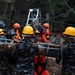 A search and rescue operation is underway at what appears to be a landslide or disaster site. Multiple rescue personnel in camouflage uniforms and hard hats are carrying a wire mesh stretcher (Stokes basket) with a victim, while a rope and pulley system is visible in the background. The muddy terrain and debris suggest a recent natural disaster such as a landslide or building collapse.