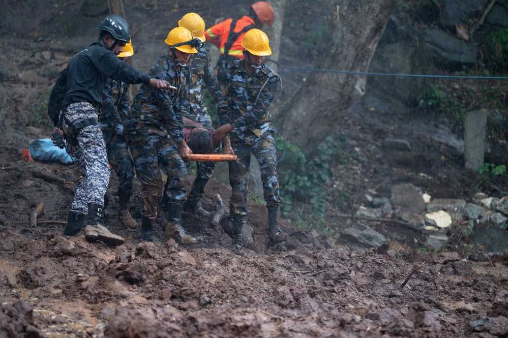 Rescue personnel in camouflage uniforms and yellow hard hats are carrying an injured or incapacitated person on a stretcher across heavily muddy, debris-strewn terrain. The scene appears to be an active disaster response operation, likely following a landslide or similar natural disaster. Additional rescue workers in orange high-visibility gear are visible in the background.