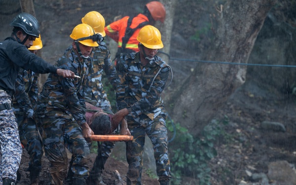 Rescue personnel in camouflage uniforms and yellow hard hats are carrying an injured or incapacitated person on a stretcher across heavily muddy, debris-strewn terrain. The scene appears to be an active disaster response operation, likely following a landslide or similar natural disaster. Additional rescue workers in orange high-visibility gear are visible in the background.