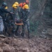Rescue personnel in camouflage uniforms and yellow hard hats are carrying an injured or incapacitated person on a stretcher across heavily muddy, debris-strewn terrain. The scene appears to be an active disaster response operation, likely following a landslide or similar natural disaster. Additional rescue workers in orange high-visibility gear are visible in the background.