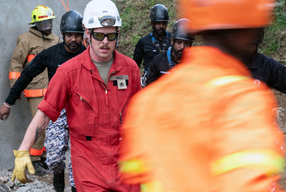 A group of emergency response personnel are gathered at what appears to be a disaster or rescue operation site. The central figure, identified by a name tag as 'HALE' with a visible rank insignia, is wearing a red jumpsuit, white Petzl helmet, and dark sunglasses. Multiple responders in varying uniforms and helmets are present, suggesting a multi-agency or multinational response effort.