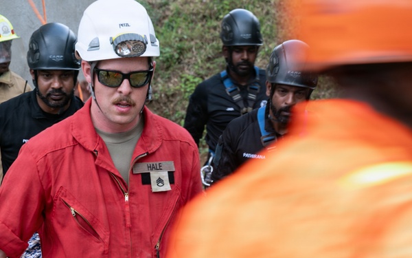 A group of emergency response personnel are gathered at what appears to be a disaster or rescue operation site. The central figure, identified by a name tag as 'HALE' with a visible rank insignia, is wearing a red jumpsuit, white Petzl helmet, and dark sunglasses. Multiple responders in varying uniforms and helmets are present, suggesting a multi-agency or multinational response effort.