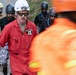 A group of emergency response personnel are gathered at what appears to be a disaster or rescue operation site. The central figure, identified by a name tag as 'HALE' with a visible rank insignia, is wearing a red jumpsuit, white Petzl helmet, and dark sunglasses. Multiple responders in varying uniforms and helmets are present, suggesting a multi-agency or multinational response effort.