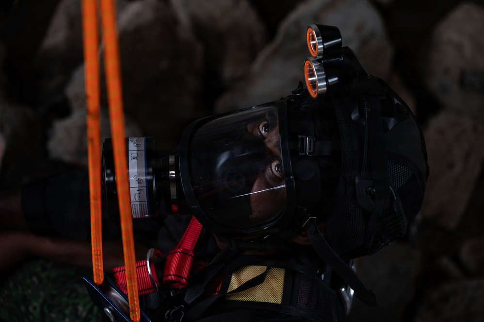 A person wearing full tactical/rescue gear including a full-face respirator mask and helmet-mounted lights is photographed in a dark environment with rocky background. The subject appears to be a first responder, rescue worker, or military personnel equipped for operations in low-visibility or hazardous conditions. Orange rope/cordage and red straps are visible in the foreground.