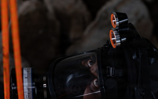 A person wearing full tactical/rescue gear including a full-face respirator mask and helmet-mounted lights is photographed in a dark environment with rocky background. The subject appears to be a first responder, rescue worker, or military personnel equipped for operations in low-visibility or hazardous conditions. Orange rope/cordage and red straps are visible in the foreground.