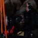 A person wearing full tactical/rescue gear including a full-face respirator mask and helmet-mounted lights is photographed in a dark environment with rocky background. The subject appears to be a first responder, rescue worker, or military personnel equipped for operations in low-visibility or hazardous conditions. Orange rope/cordage and red straps are visible in the foreground.