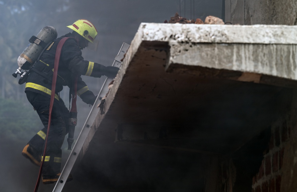 A firefighter in full protective gear is climbing a metal ladder toward the roof or upper level of a damaged concrete structure surrounded by heavy smoke. The firefighter is equipped with a self-contained breathing apparatus (SCBA) tank on their back and is carrying a fire hose. The scene depicts an active emergency response operation in a smoke-filled environment.