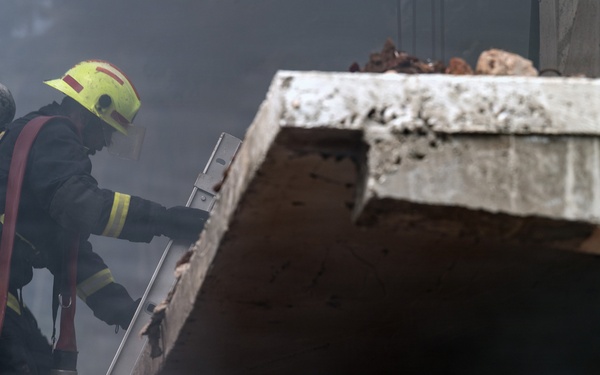 A firefighter in full protective gear is climbing a metal ladder toward the roof or upper level of a damaged concrete structure surrounded by heavy smoke. The firefighter is equipped with a self-contained breathing apparatus (SCBA) tank on their back and is carrying a fire hose. The scene depicts an active emergency response operation in a smoke-filled environment.